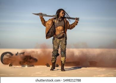 Post-apocalyptic biker woman with weapon outdoors. Young slim girl warrior in shabby clothes holding sword standing in a confident pose against the broken burning motorcycle looking away. Nuclear post