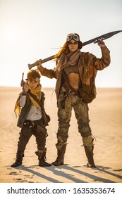Post-apocalyptic woman and boy with weapons outdoors. Desert and dead wasteland on the background. Aggressive girl warrior in shabby clothes holding the sword and young boy with a gun standing in a