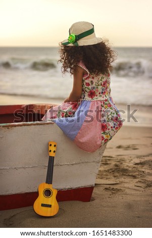 Postcard of girl sitting with hat in a boat on the beach with a guitar