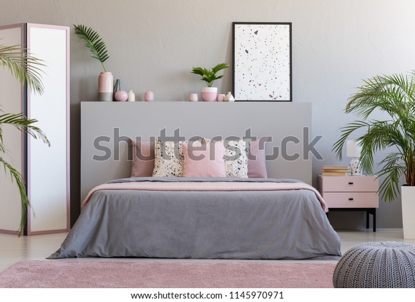 Poster on grey headboard of bed in\
grey and pink bedroom interior with plants. Real\
photo