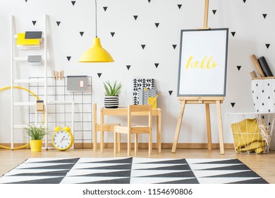 Poster On A Wooden Easel And A Small Desk For Children In A White, Scandi Style Preschool Room Interior With White Wall And Vibrant, Yellow Decorations