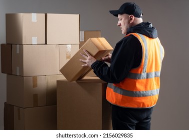 Postman Looks At Box. Postman In Orange Vest On Gray. Postal Service Worker With Boxes. Mature Postman Back To Camera. Career In Post Office Concept. Man In Black Baseball Cap In Warehouse