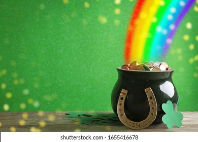 Pot with gold coins, horseshoe and clover leaves on wooden table against green background, space for text. St. Patrick's Day celebration