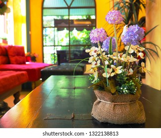 A Pot Put On Wooden Table In Yellow Room. Flowers Color Are Violet, Purple, And White With Green Leaves. Red Sofa In The Room. No People. A Room Is Boutique. 