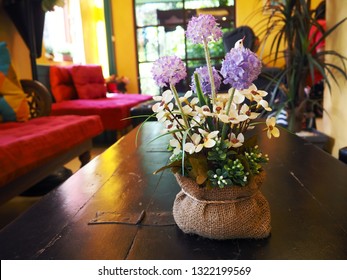 A Pot Put On Wooden Table In Yellow Room. Flowers Color Are Violet, Purple, And White With Green Leaves. Red Sofa In The Room. No People. A Room Is Boutique. 