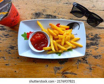 Potato French Fries Served With Spicy Chilly And Tomato Sauce On The Wooden Table. French Fries, Black Sunglasses, And Red Velvet Drink. 