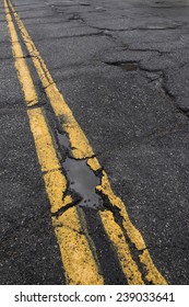 Pothole And Cracked Road With Yellow Dividing Line  In Need Of Repair.