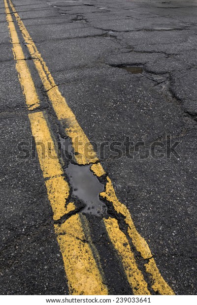 Pothole and cracked road with yellow dividing\
line  in need of\
repair.