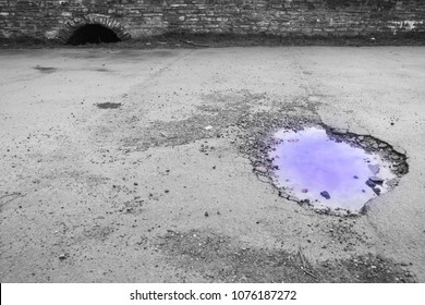 Pothole In UK Road With Bright Blue Water And Sewer Entry Archway In Background