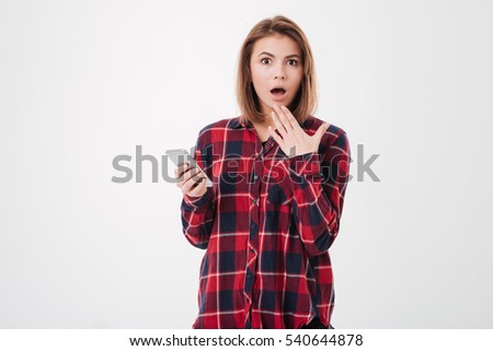 Potrait of an astonished young woman in plaid shirt holding mobile phone and looking at camera over white background