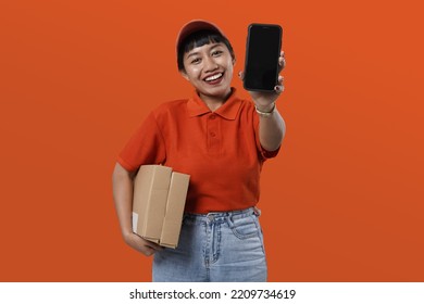 Potrait of young asian courrier woman wearing a red cap and uniform while holding a smartphone and a brown package. Female delivery courier working for a delivery mobile app