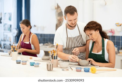 In pottery workshop, apprentice female students sitting at table, engaged in making products from raw clay in presence of teacher. Visitors to clay craft workshop work under guidance of male mentor - Powered by Shutterstock