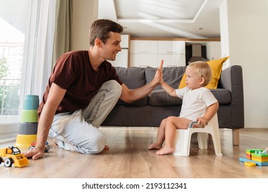 Potty Training. Young Father Assisting His Little Baby Boy Using Pot In Living Room. Cute Baby Giving High Five To His Dad Sitting On Potty At Home. Dad Teaching His Son How To Use Potty