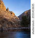 The Poudre River in early winter.  Flowing stream with ice in foreground and late afternoon sunlit hillside under a blue sky.  Dark river water with pockets of ice with river rock. 