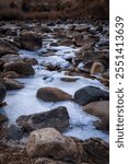 The Poudre River in early winter.  Flowing stream with ice in foreground and late afternoon sunlit hillside under a blue sky.  Dark river water with pockets of ice with river rock. 