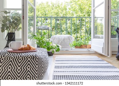 Pouf with pink plate with coffee mug and croissant in blurred foreground in real photo of room with fresh plants, rug on the floor and open door to balcony