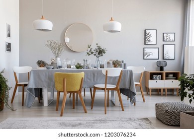 Pouf, Plants And Round Mirror In Bright Dining Room Interior With Chairs At Table Under Lamps. Real Photo