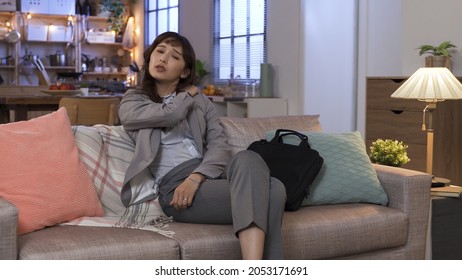 Pounding On Sore Neck After Long Tiring Day. Korean Businesswoman Approaching Living Room Sofa And Sitting Down With Exhaustion. Genuine Lifestyle