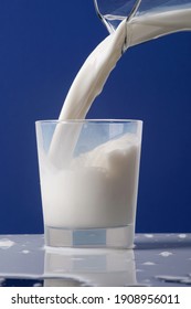 Pour The Creamy Milk From The Carafe Or Jug Into A Glass. The Milk Spills Over The Table. Photo Isolated On Blue Background