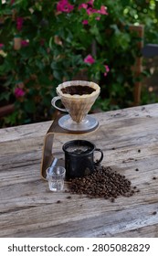 Pour over coffee dripping through a filter into a black mug beside scattered coffee beans on a rustic wooden table with tropical greenery, artisan brewing process, detail shot