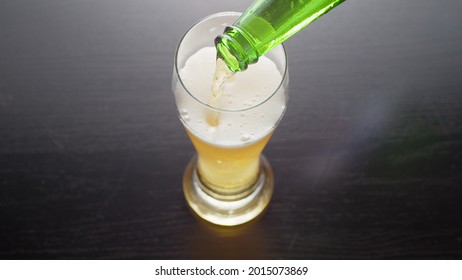 Pouring Beer Into Glass From Bottle, View From Above. Half Pint Of Frothy Fresh Lager Beer On Table. Top View.