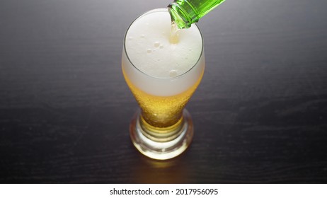 Pouring Beer Into Glass From Bottle, View From Above. Half Pint Of Frothy Fresh Lager Beer On Table. Top View.