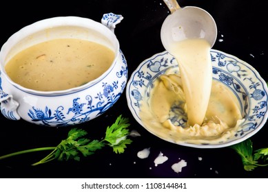 Pouring Cauliflower Soup In To Festive Plate With Ladle, Porcelain Bowl, Parsley Leaf, On Spilled Black Background.