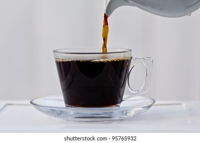 Pouring Coffee Into An Almost Full Clear Cup With A Saucer.