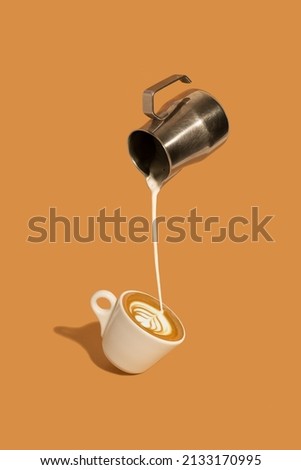 Pouring creamy milk from a stainless steel milk jug, and making late art espresso in a coffee cup on brown background.