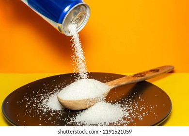 Pouring Crystal Sugar From A Blue Can To A Wooden Spoon On A Black Plate And Orange Background.  Motion Blur On Sugar Texture. Sugar Danger Concept