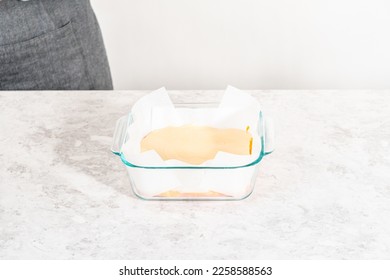 Pouring fudge mixture into the square glass baking pan lined with parchment paper to prepare candy corn fudge. - Shutterstock ID 2258588563