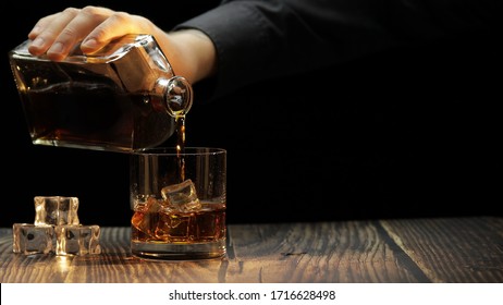 Pouring Of Golden Whiskey, Cognac Or Brandy From The Bottle Into Glass With Ice Cubes On Wooden Table. Black Background. Side View Of Bartender Pouring Glass Of Alcohol Drink Bourbon, Rum