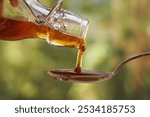 Pouring herbal syrup from a glass bottle onto a metal spoon, closeup