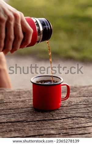 Pouring hot steaming tea into bright red enamel coffee mug staying on old log in nature. Selective focus on mug rim.