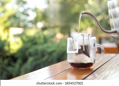 Pouring hot water into glass cup with drip coffee bag from kettle on wooden table. Space for text
