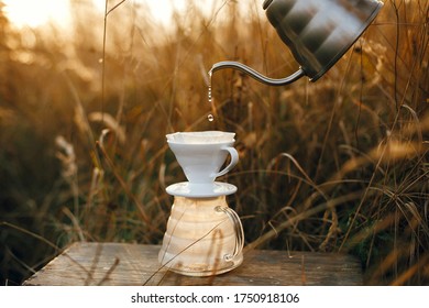 Pouring hot water from steel kettle in glass flask dripper with filter on background of rural countryside and herbs in sunset. Alternative coffee brewing outdoors. Atmospheric rustic moment - Powered by Shutterstock