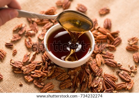 Pouring Maple Syrup from Spoon. a close up view of a bowl of maple syrup with a spoon dipped in and pouring some out surrounded by pecans on a textured brown cloth
