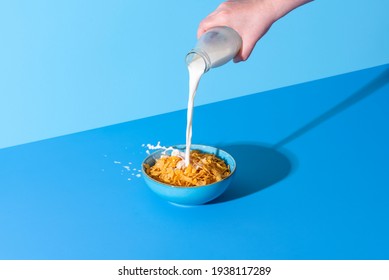 Pouring milk from a bottle into a bowl with cornflakes cereals. Bowl with corn cereals and milk isolated on a blue background - Shutterstock ID 1938117289