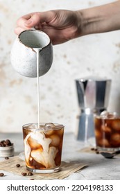 Pouring milk in coffee with ice cubes with beautiful twists on light background. Vertical orientation. Iced latte coffee still life