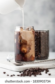 Pouring Milk Into Glass With Iced Black Coffee With Glass Container Of Beans And Spoon On Marbel Board And Light Table Background.