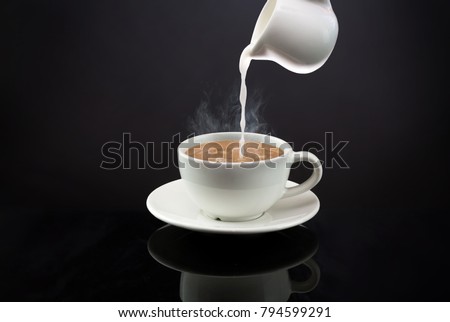 Pouring a milk in to a mug of hot coffee or tea on black background