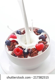 Pouring Milk With A Splash Into A Bowl Of Crunchy Breakfast Cereal Topped With Fresh Berries With Raspberries And Blueberries Over White With Copyspace