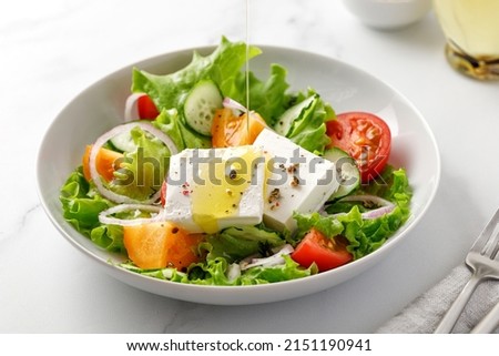 Pouring olive oil on greek salad with green leaves, tomatoes, cucumber, feta cheese. White bowl, marble background