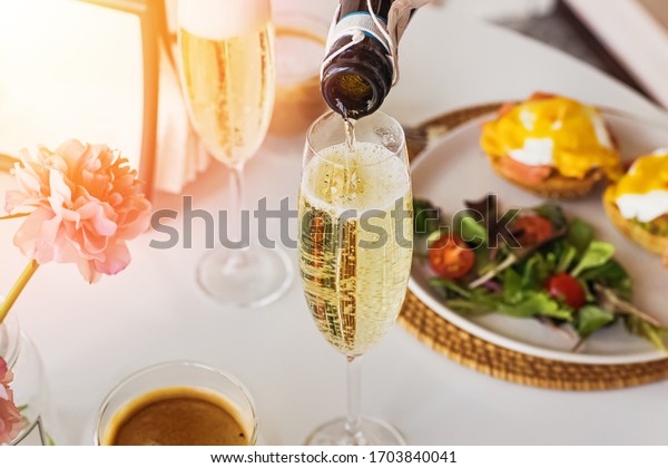 Pouring prosecco sparkling\
wine in a glass on sunday brunch with blurred eggs royal on the\
background.