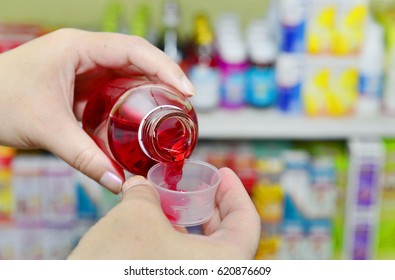 Pouring Red Cough And Cold Liquid Medicine Into Measuring Cup On Pharmacy Drugstore Background.