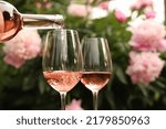 Pouring rose wine from bottle into glass against beautiful peonies, closeup