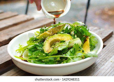 Pouring soy sauce dressing in plate of salad consists sliced grilled avocado, coriander, lettuce and sesame seeds, close up healthy organic diet food on wooden table