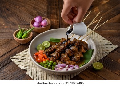 Pouring Soy Sauce On Sate Daging Sapi Bumbu Ketumbar Or Coriander Beef Satay, Served On Plate With Onion, Chilli, Soy Sauce And Tomato. Indonesian Traditional Food Verry Popular During Eid Al Adha