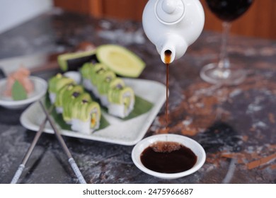 pouring soy sauce from a small ceramic jug into a small white bowl, with a plate of sushi rolls in the background - Powered by Shutterstock