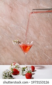 Pouring strawberry alcoholic or non-alcohol cocktail from glass bottle into martini glass, decorated with blooming branches of cherry tree over white and pink texture background - Shutterstock ID 1973153126